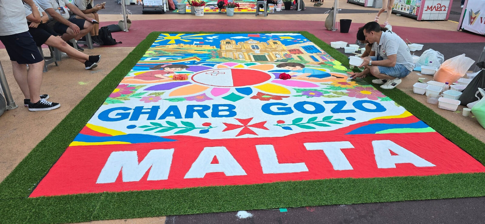 A colourful sand carpet reading Għarb Gozo Malta, with the Maltese flag and a Maltese cross, as volunteers add final details