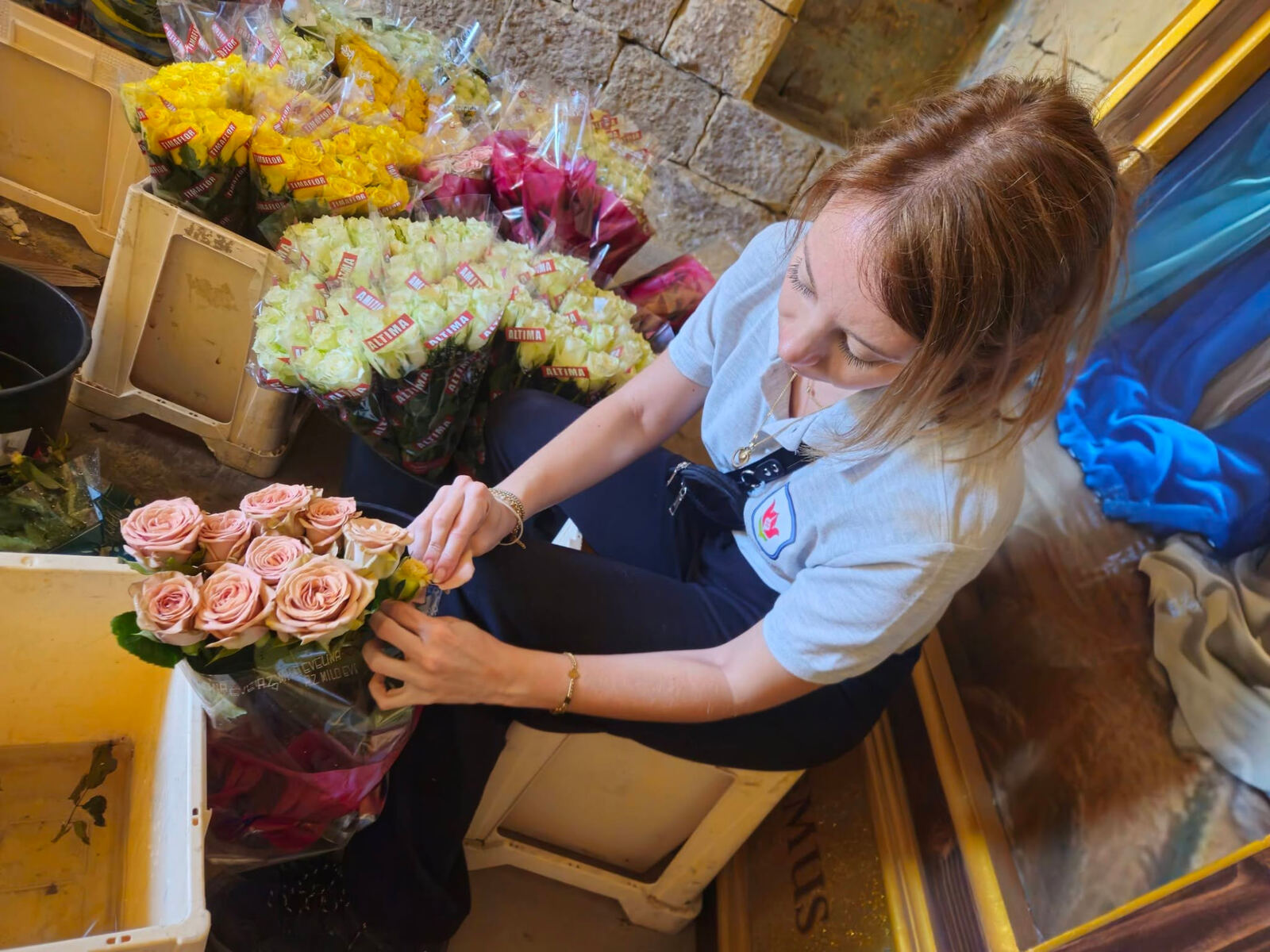 A volunteer seated among crates of roses, carefully preparing a bunch of pale pink roses